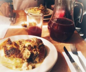 Im Vordergrund ein Teller mit Kartoffelgratin und gefüllter Paprika, daneben Besteck auf einer Serviette. Im Hintergrund stehen eine Glaskaraffe und ein Glas, die mit einer rötlich-lila dunklen Flüssigkeit gefüllt sind. Noch weiter im Hintergrund ist ein weiterer Teller mit der gleichen Mahlzeit zu sehen.